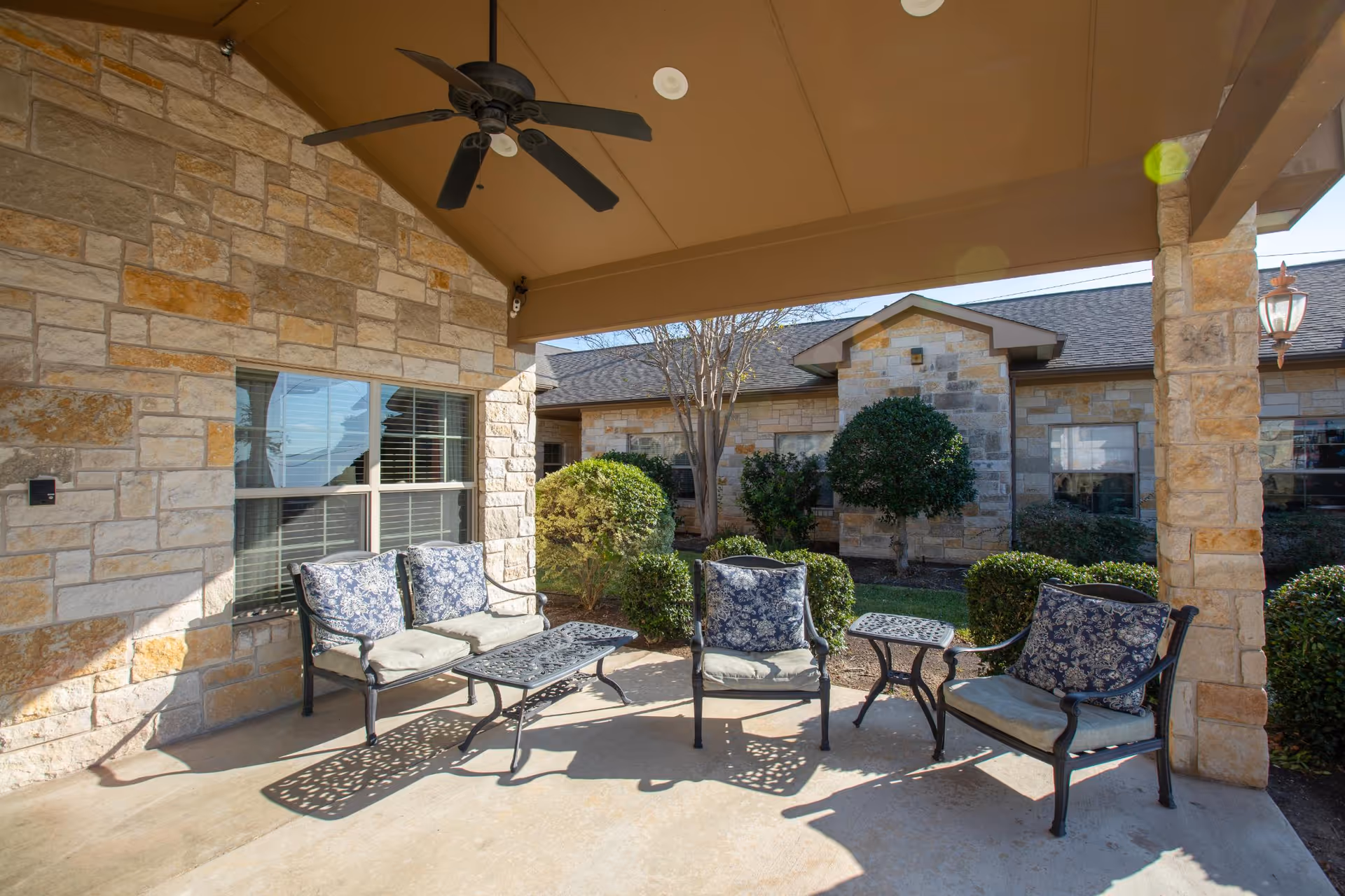Covered outdoor patio area with stone walls and ceiling fan, featuring cushioned metal chairs and a loveseat with floral patterned cushions, a coffee table, and a small side table. The patio overlooks a landscaped garden with bushes and trees, and the exterior of the building is visible in the background.
