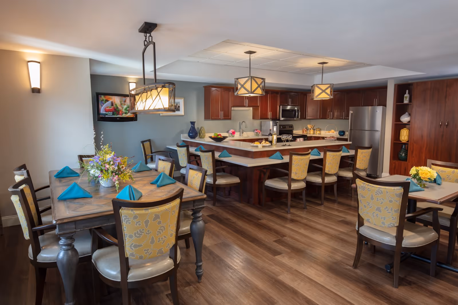 A well-lit dining area with wooden floors featuring a large wooden dining table set with teal napkins and floral centerpiece, surrounded by chairs with yellow floral upholstery. In the background, there is a kitchen area with dark wood cabinets, stainless steel appliances, and a countertop with bar stools. Pendant lights hang above the kitchen island, and a wall-mounted TV is visible on the left wall.