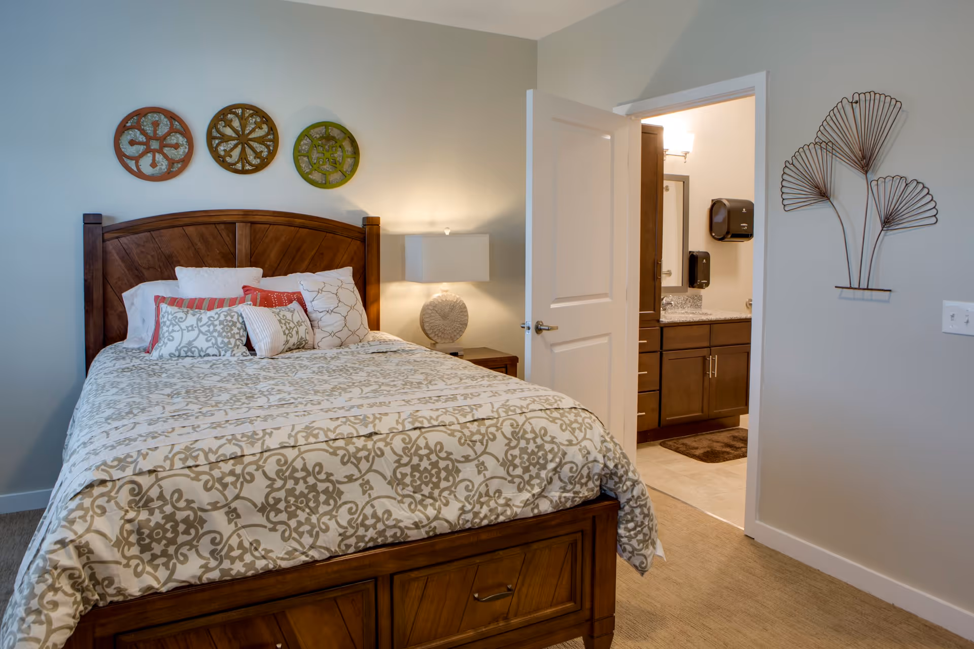 A bedroom with a wooden bed frame and a patterned bedspread. The bed is adorned with multiple decorative pillows in white, red, and patterned designs. Above the bed, three circular decorative wall hangings in red, brown, and green are displayed. To the right of the bed is a nightstand with a lamp. An open door reveals a bathroom with a granite countertop, wooden cabinets, and a wall-mounted soap dispenser. On the wall to the right of the door, there is a metal wall art piece shaped like abstract leaves.
