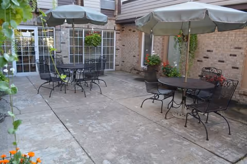 Outdoor patio area with two round metal tables, each with four matching chairs and large umbrellas providing shade. The patio is surrounded by brick walls with windows and doors, and there are hanging flower baskets and potted plants adding greenery and color.