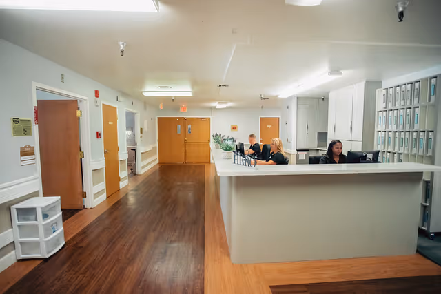 Reception area inside a senior living facility with a long white counter where three staff members are seated working on computers. The floor is wooden, and there are multiple doors along the left wall. Filing cabinets and shelves are visible behind the reception desk.