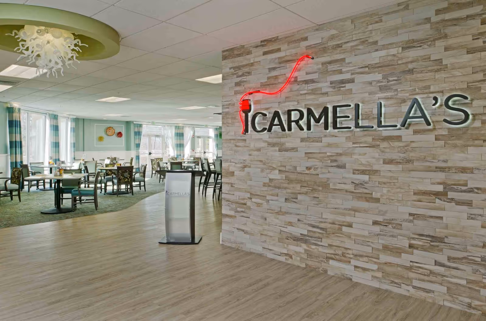 Interior view of Carmella's dining area at Lorien Harmony Hall, featuring a modern design with a stone accent wall displaying the Carmella's sign with a red neon fork, wooden flooring, multiple dining tables and chairs, large windows with curtains allowing natural light, and a decorative ceiling light fixture.
