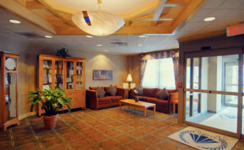 A cozy living room area in a senior living facility featuring two brown sofas with cushions, a wooden coffee table, a wooden cabinet with glass doors, a large potted plant, a grandfather clock, a ceiling light fixture, and a glass entrance door with an exit sign above it. The room has patterned carpet flooring and a window with curtains allowing natural light in.