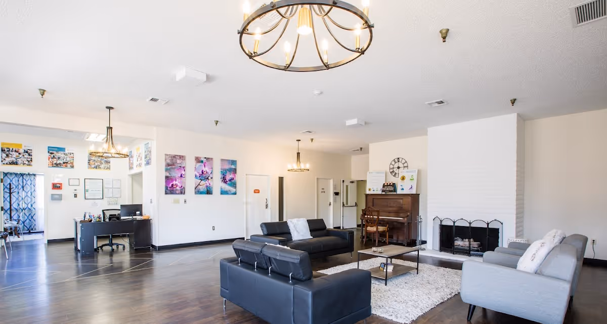 A spacious living room area with modern black and gray sofas arranged around a coffee table on a white rug. There is a white brick fireplace with a black metal screen, a wooden piano with a chair, and a large wall clock above it. The room has wooden flooring, white walls, and two hanging light fixtures. In the background, there is a desk with a computer and some colorful artwork on the walls.