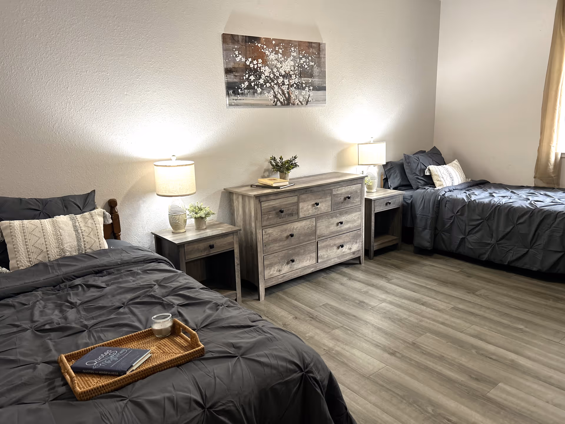 A bedroom with two twin beds featuring dark gray bedding and decorative pillows. Between the beds is a wooden dresser with six drawers, flanked by two matching nightstands each holding a lamp and a small plant. A framed artwork of white flowers hangs on the wall above the dresser. The room has light-colored walls and wood flooring, with a window covered by a beige curtain on the right side.