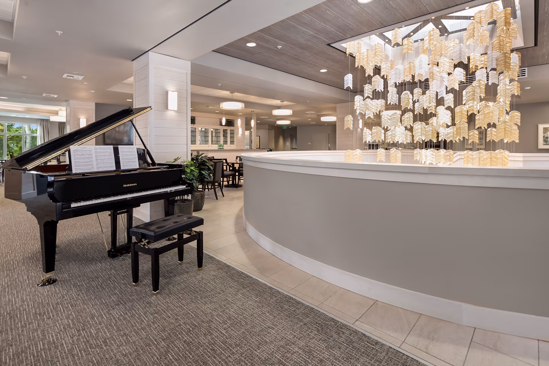 Interior view of a senior living facility featuring a black grand piano with sheet music on the stand and a matching bench. To the right, there is a curved reception desk with a modern hanging light fixture above it. In the background, there are dining tables and chairs, light fixtures on the ceiling, and large windows letting in natural light.