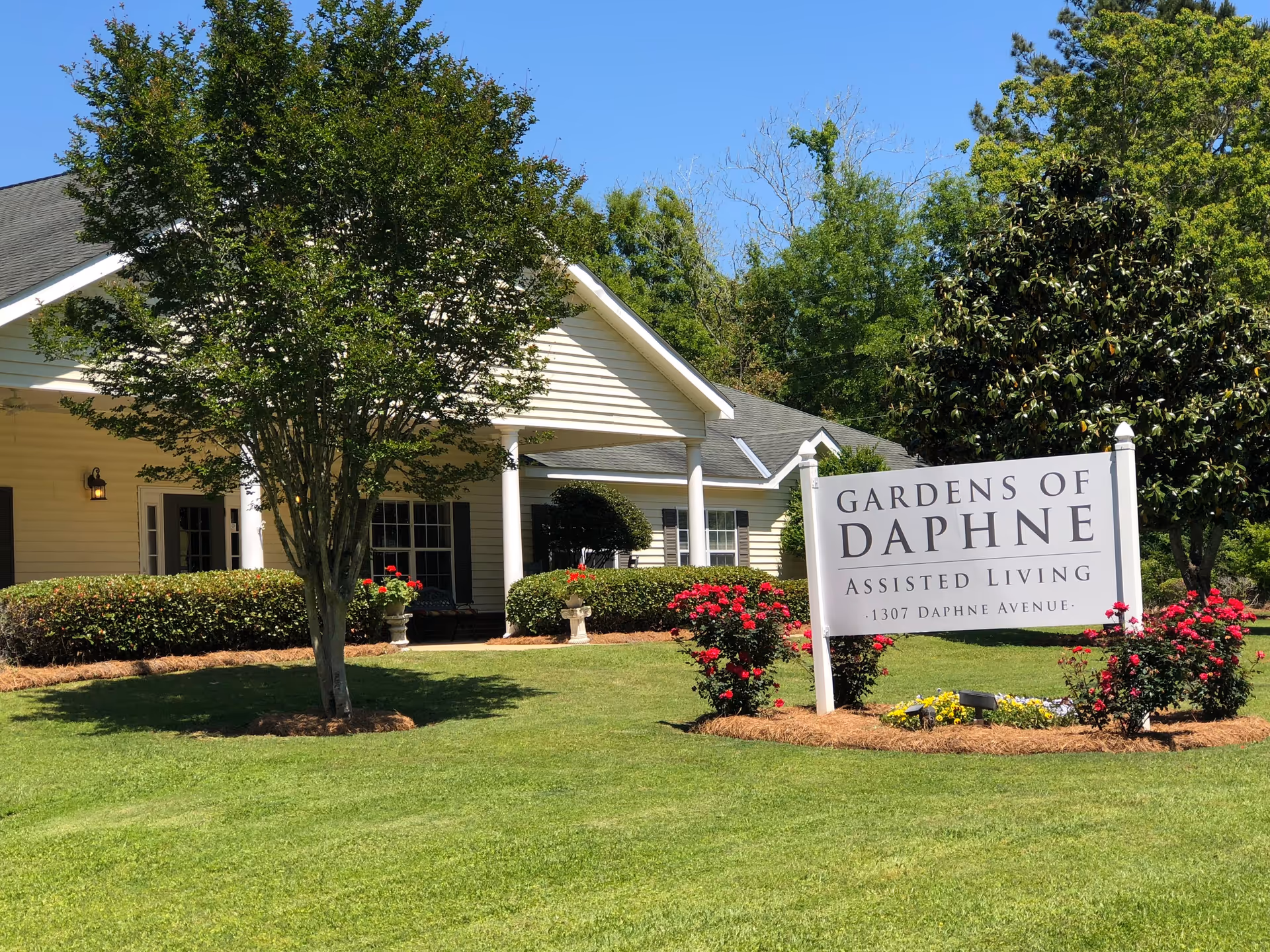 Exterior view of Gardens of Daphne assisted living facility with a white building, green lawn, trees, and a sign displaying the facility name and address.