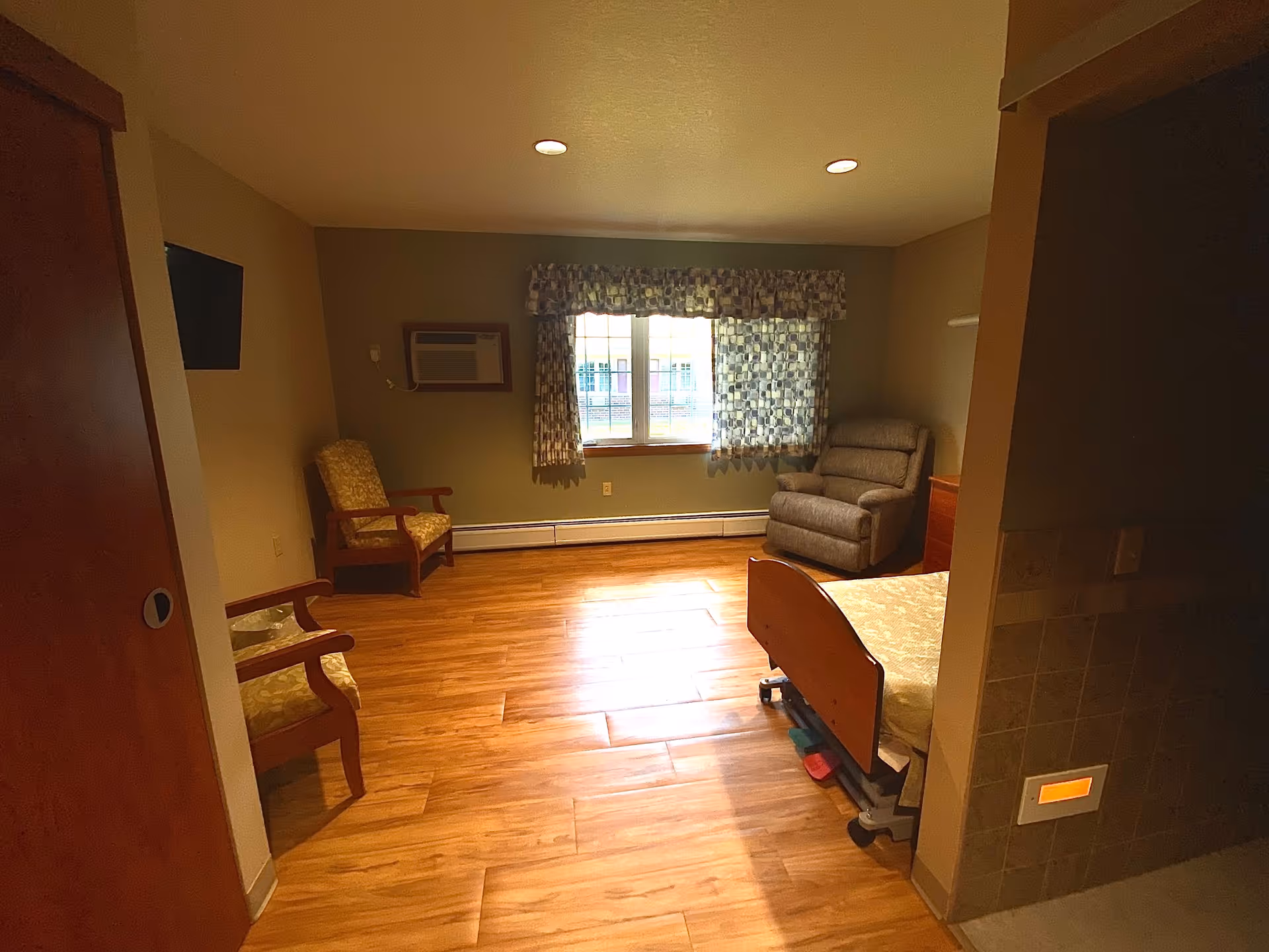 A senior living facility room with wooden flooring, two armchairs with patterned upholstery, a recliner chair near a window with patterned curtains, a hospital-style bed with a wooden headboard, and an air conditioning unit mounted on the wall. The room has warm lighting and neutral-colored walls.