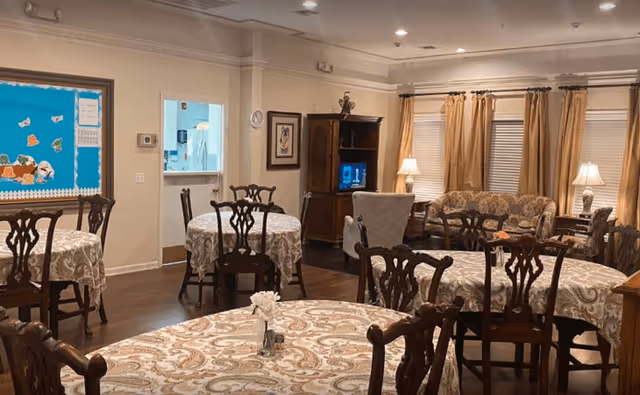 A cozy dining area in a senior living facility with several round tables covered with patterned tablecloths and wooden chairs. In the background, there is a seating area with a sofa, armchairs, and lamps near windows with beige curtains. A wooden cabinet with a television is also visible, along with a bulletin board on the wall and a doorway leading to another room.