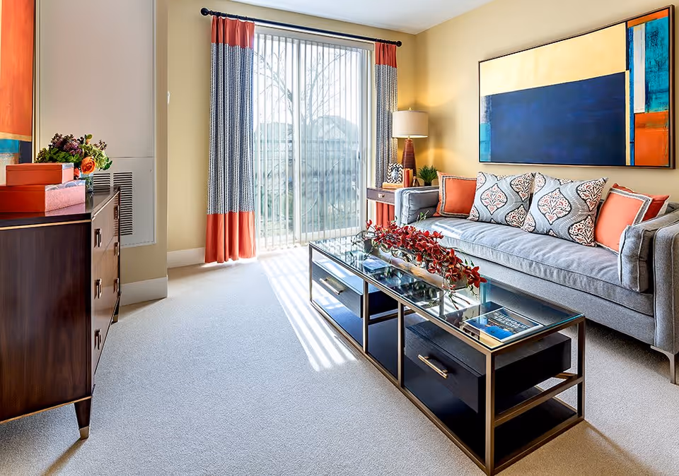 A bright living room with a gray sofa adorned with patterned and solid orange cushions, a glass coffee table with red floral decoration, a wooden sideboard with decorative items, and a large abstract painting on the wall. The room has beige walls, carpeted floor, and a sliding glass door with curtains letting in natural light.