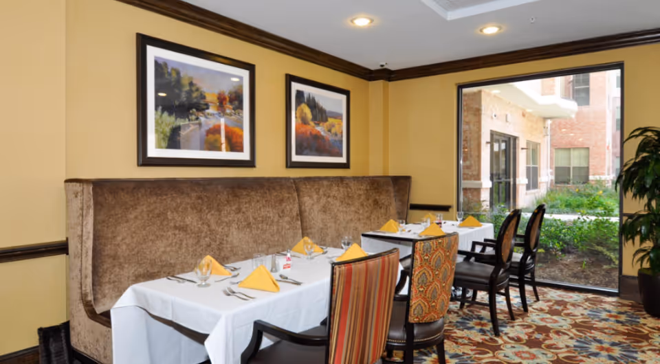 Dining room with a table set with yellow napkins, upholstered banquette seating, patterned chairs, framed artwork, and a large window overlooking a courtyard.