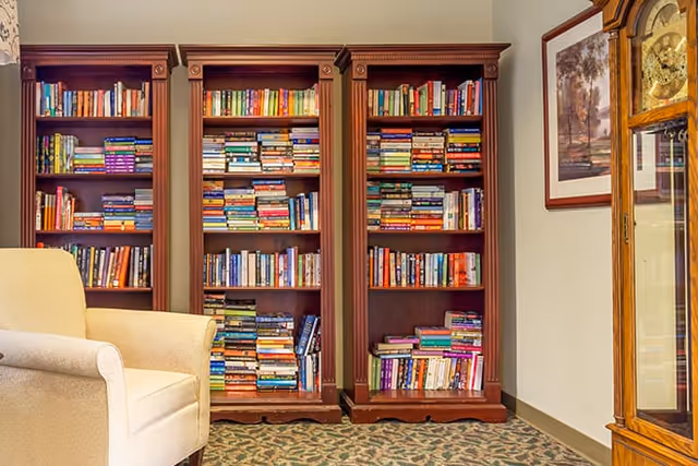 A cozy reading area featuring three tall wooden bookshelves filled with a variety of books. To the left, there is a beige upholstered armchair. On the right side, part of a wooden grandfather clock and a framed picture on the wall are visible. The floor is carpeted with a patterned design.