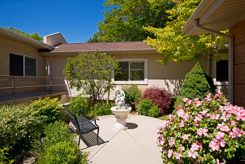 A sunny outdoor garden area at Crestwood Rehabilitation and Nursing featuring a concrete pathway, a black metal bench, a white decorative fountain with a statue, and various green shrubs and flowering plants including pink flowers. The garden is surrounded by building walls with windows and a clear blue sky overhead.