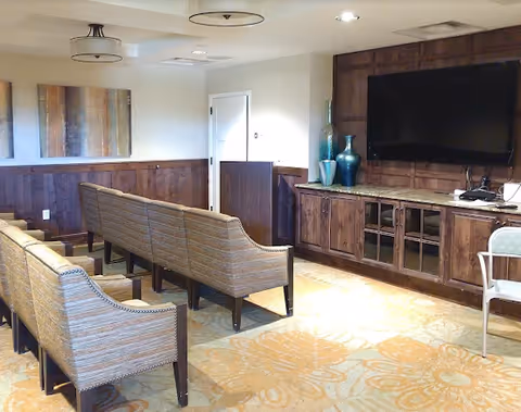 A cozy living room area with two rows of upholstered chairs facing a large flat-screen TV mounted on a wooden paneled wall. Below the TV is a wooden cabinet with glass doors and decorative vases on top. The room has a patterned carpet and neutral-colored walls with two abstract paintings hanging on one side.