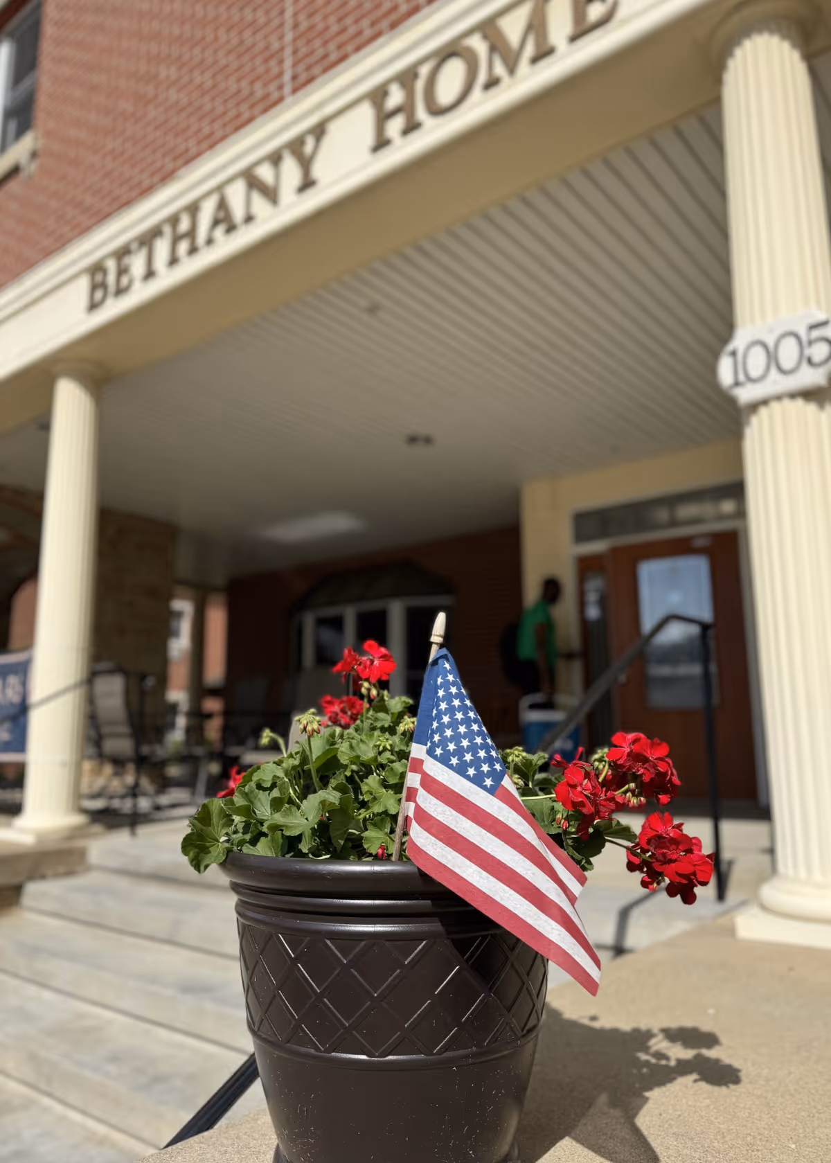 Potted red geraniums with a small American flag in front of the covered entrance of Bethany Home Retirement Center.