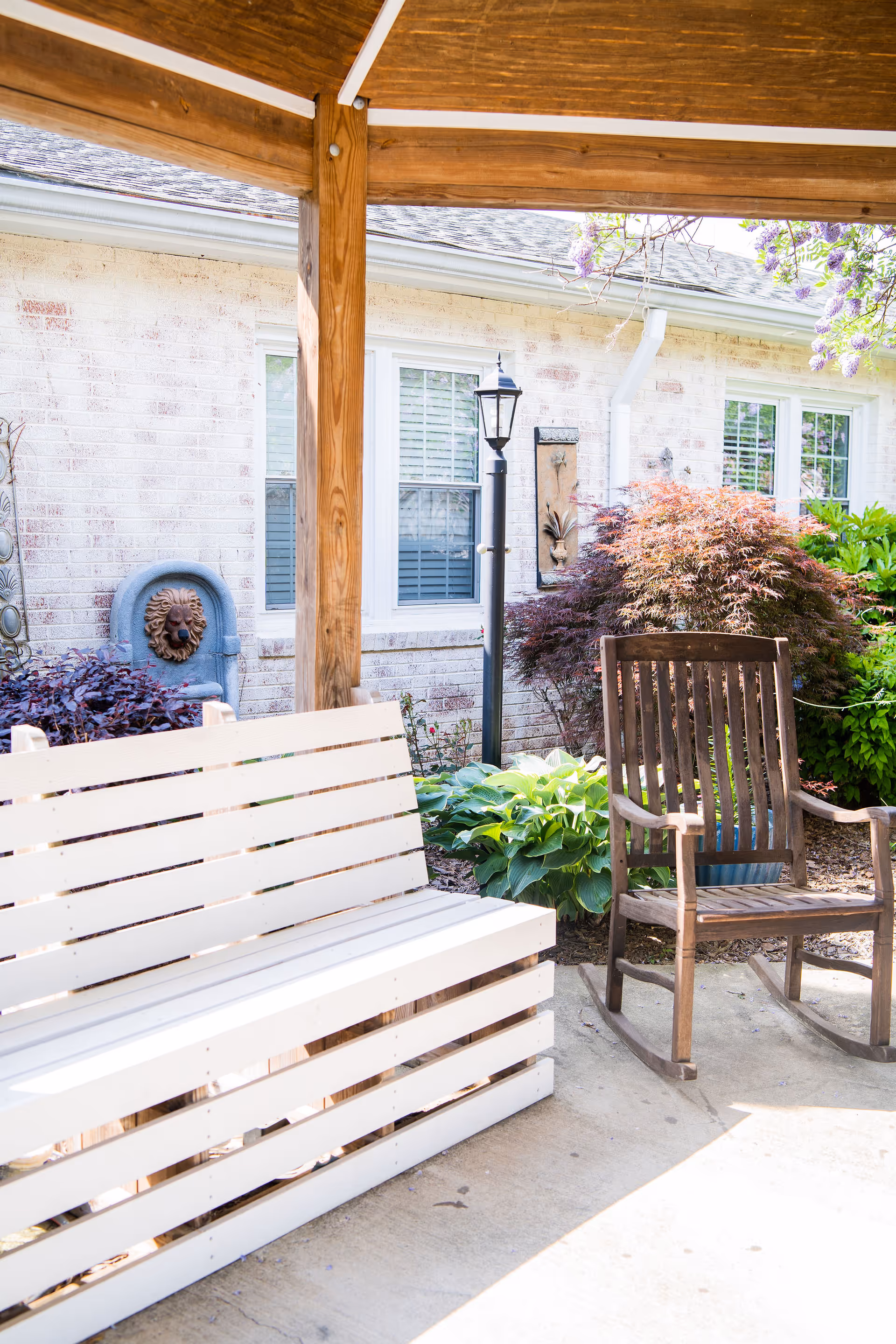 Outdoor patio with a white wooden bench, a wooden rocking chair, potted plants, and a brick building facade under a wooden pergola.