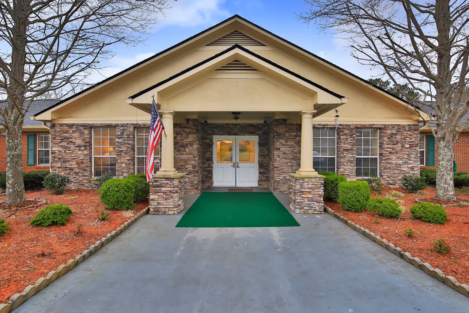 Front entrance of a single-story building with stone and brick exterior, two columns supporting a covered porch, an American flag mounted on the left column, and a green mat leading to double white doors. There are landscaped bushes and trees on either side of the entrance.
