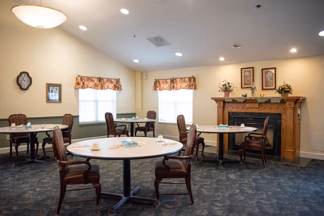 Communal dining room with round tables and chairs, windows with valances, and a decorative fireplace.