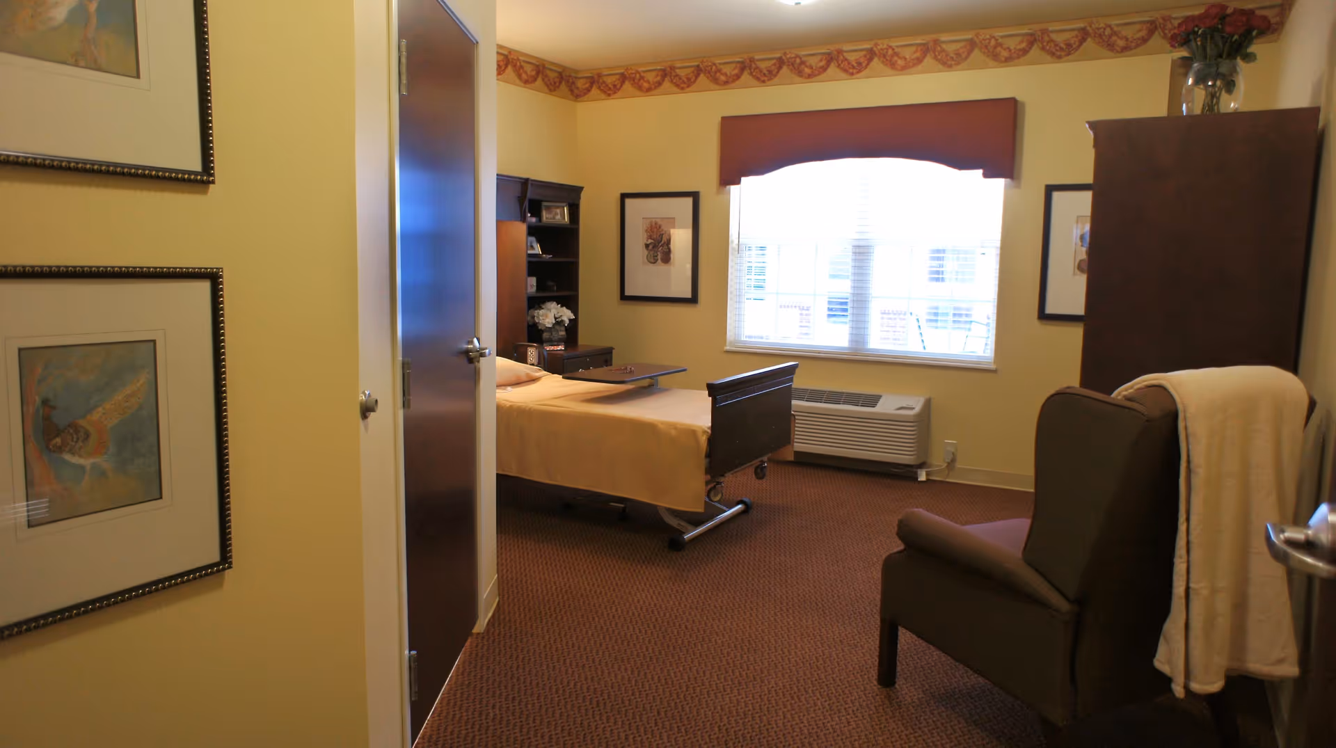 A cozy bedroom in a senior living facility with a single bed covered in a beige blanket, a dark wooden bookshelf and cabinet, a window with blinds and a red valance, a brown armchair with a white blanket draped over it, and framed artwork on the yellow walls.