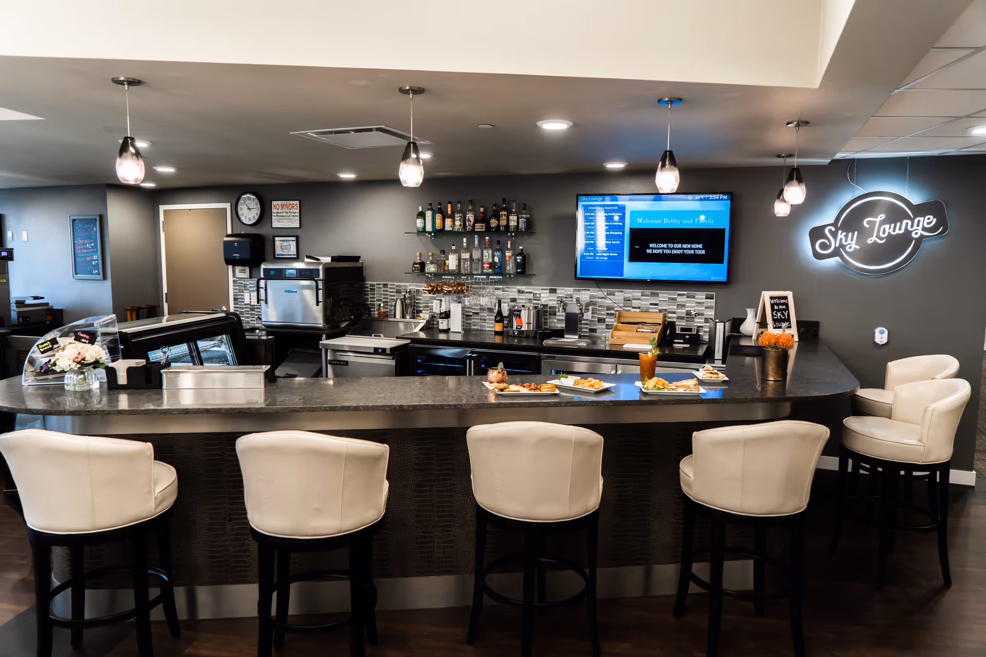 Interior view of a modern lounge bar area named Sky Lounge with a curved counter, five white cushioned bar stools, hanging pendant lights, a variety of liquor bottles on shelves, a digital screen displaying a welcome message, and plates of food and drinks on the counter.