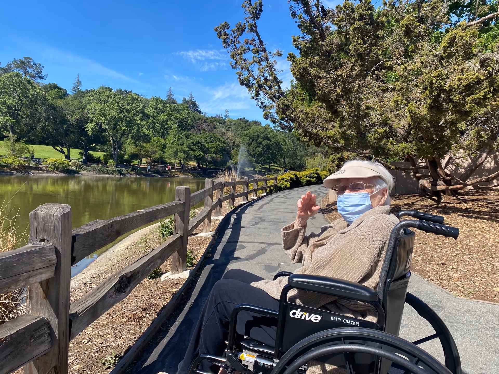 An elderly woman wearing a beige sweater, visor, glasses, and a face mask is sitting in a wheelchair on a paved path next to a wooden fence. The path runs alongside a pond with green trees and a clear blue sky in the background.