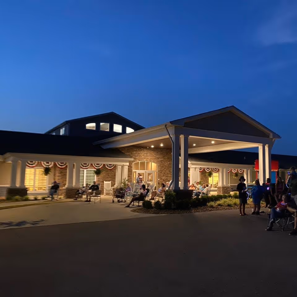 Evening view of the entrance to Colonial Senior Living facility with people sitting and standing outside under a covered porch decorated with patriotic bunting.