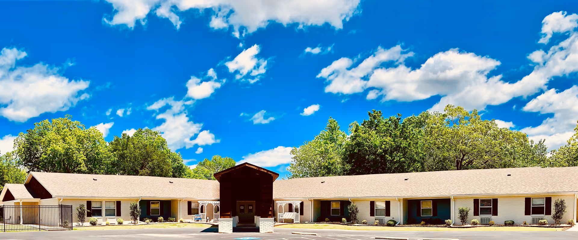 Exterior view of a single-story senior living facility building with a light-colored roof and walls, surrounded by green trees under a bright blue sky with scattered clouds. The building has multiple windows and a central entrance with a covered porch.