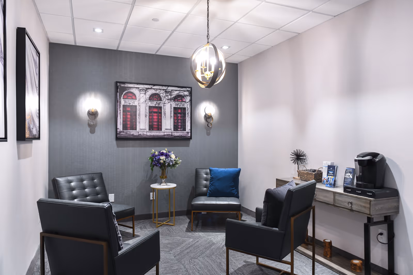 A small, modern sitting area with four black leather chairs arranged around a small round table with a vase of flowers. The walls are light gray with a darker gray accent wall featuring a framed picture of a building facade with red windows. A pendant light hangs from the ceiling, and a console table against one wall holds a coffee maker and informational brochures.
