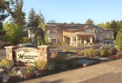 Exterior view of Hearthstone of Beaverton facility showing a two-story building with a covered entrance, surrounded by trees, shrubs, and a landscaped garden with flowers. A sign with the facility name is visible near the sidewalk.