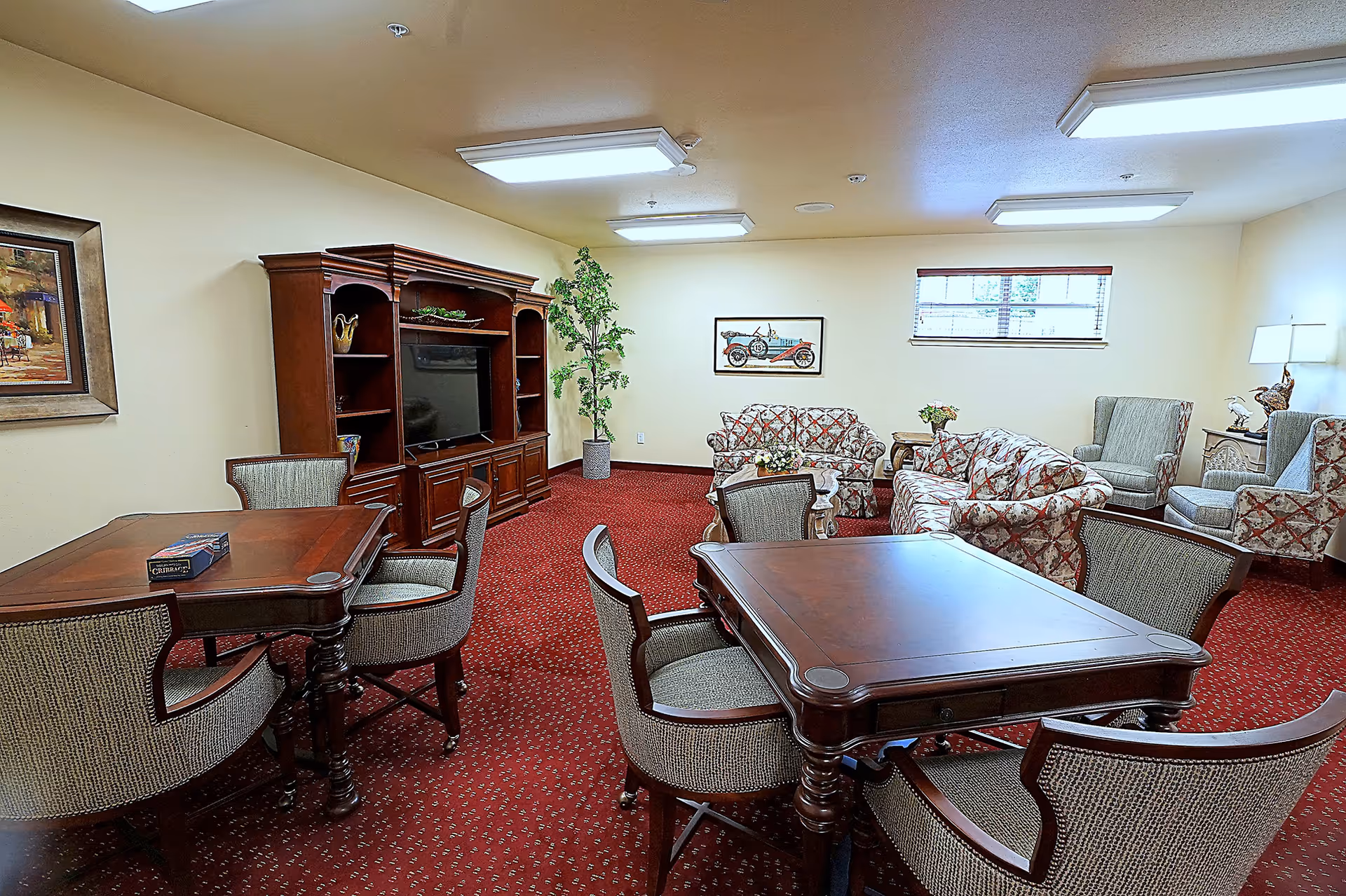 Shared sitting room with wooden tables and upholstered chairs, patterned sofas, a TV cabinet, and red carpet.