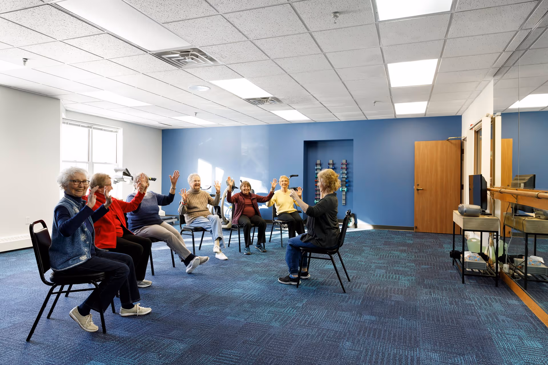 A group of elderly individuals seated in a spacious, well-lit room with blue carpet and a blue accent wall, participating in a seated exercise or stretching activity led by an instructor. The room has a large mirror on one side and exercise equipment on a rack against the wall.
