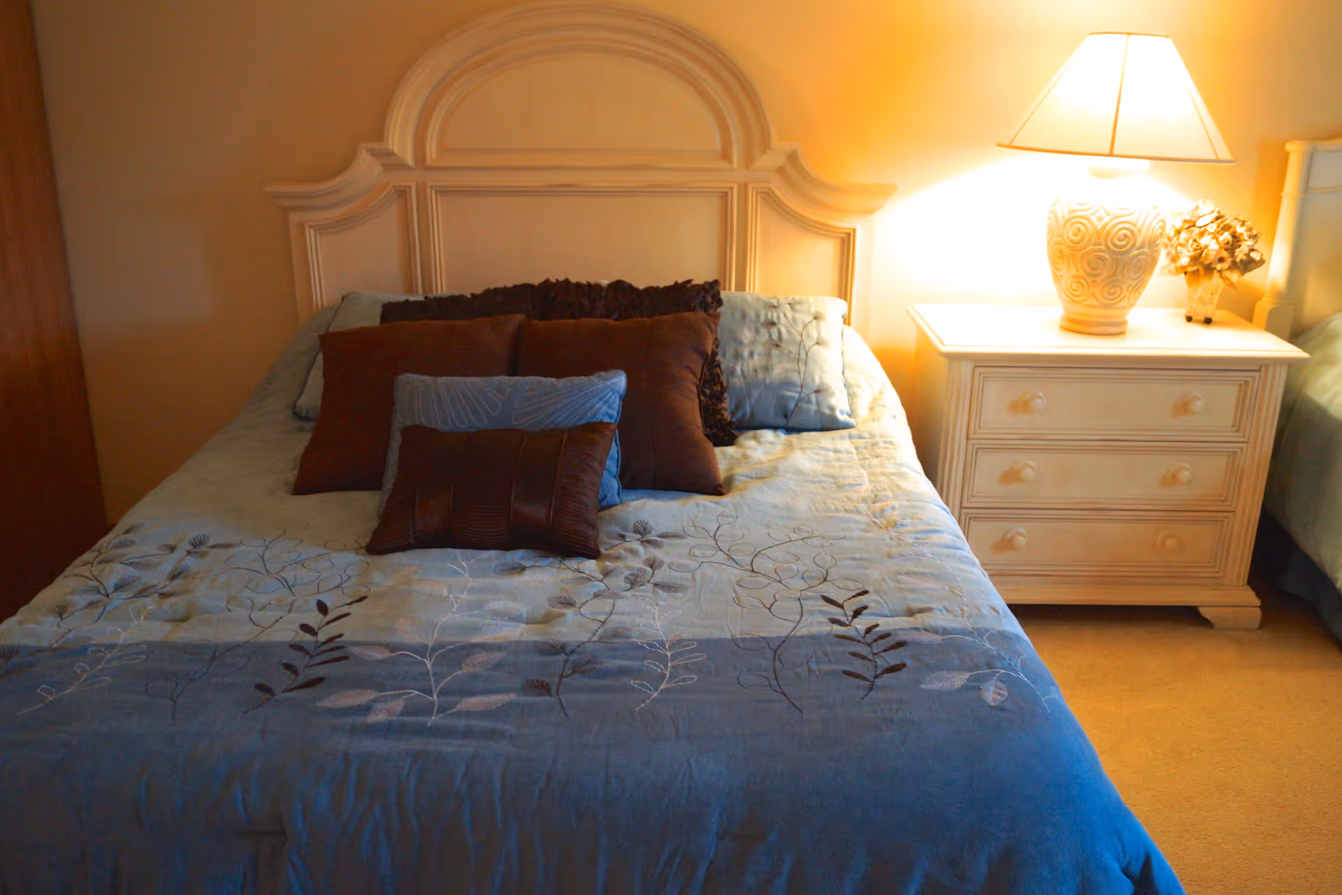 A neatly made bed with a decorative headboard, covered with a blue and beige floral-patterned bedspread and several brown and blue pillows. To the right of the bed is a white nightstand with three drawers, a decorative lamp, and a small flower arrangement. The room is softly lit, creating a warm and cozy atmosphere.