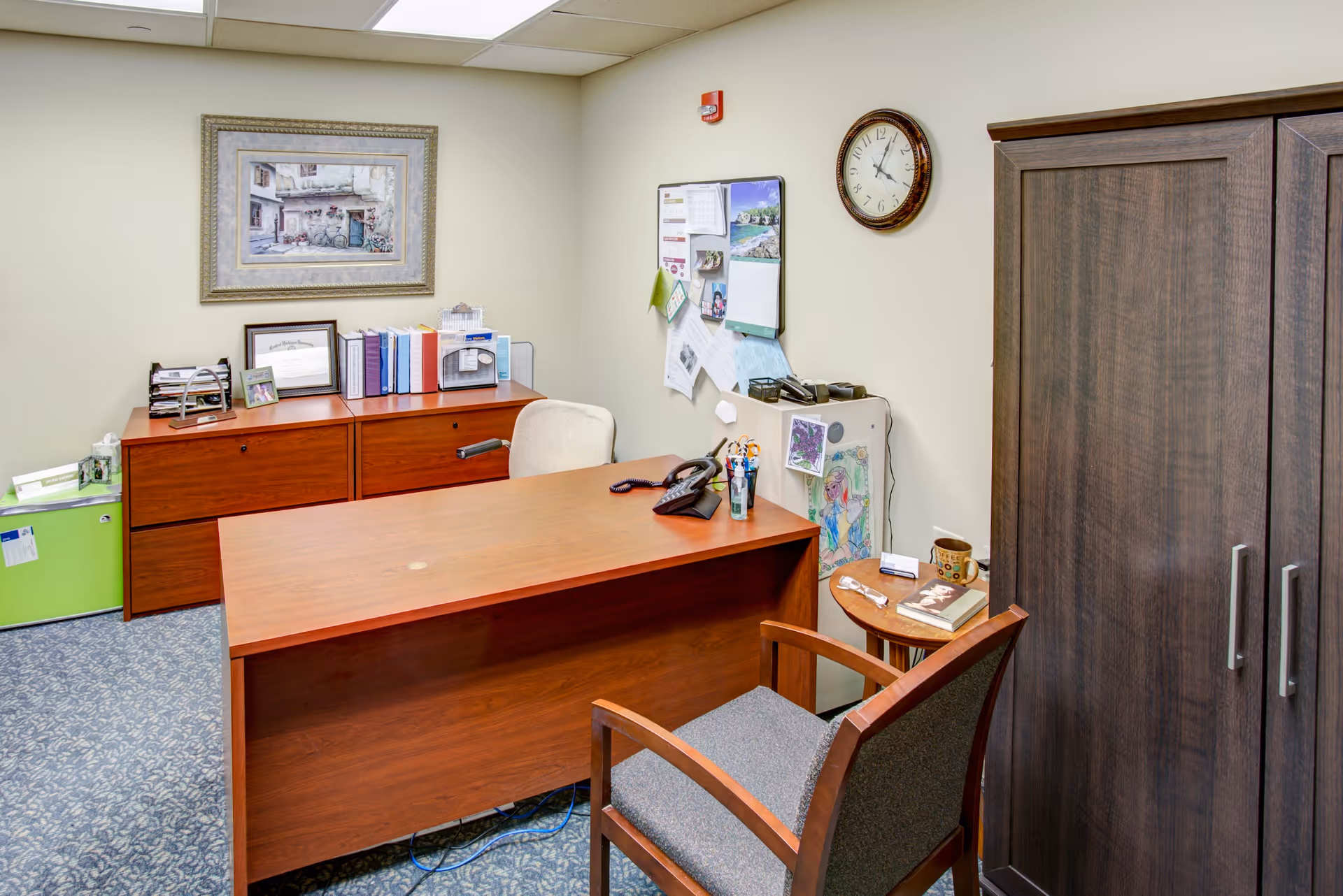 Private office with a wooden desk, guest chair, filing cabinets, bulletin board, and wall clock.