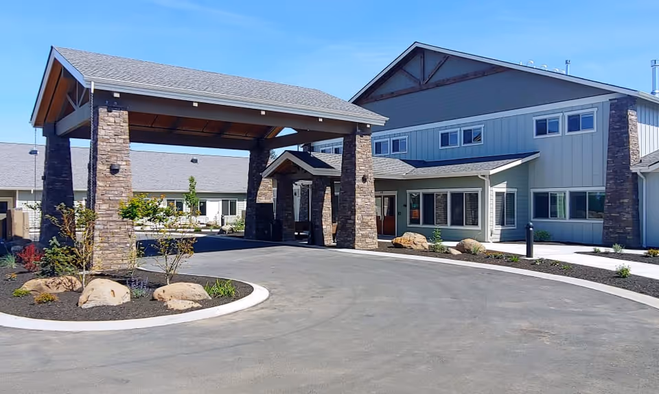 Exterior view of The Lodge In Sisters senior living facility showing a covered entrance with stone pillars, a driveway, and landscaped areas with small plants and rocks under a clear blue sky.