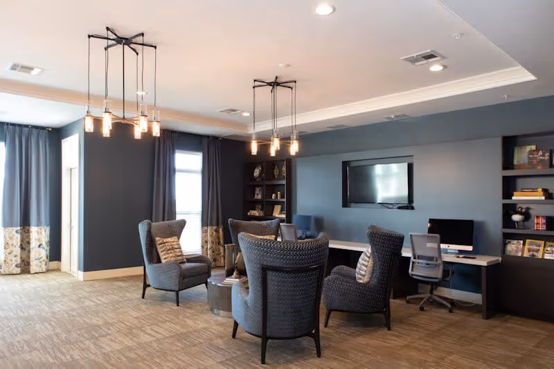 A cozy senior living common area with four gray upholstered armchairs arranged around a small round table. The room features blue walls, two windows with curtains, built-in shelves with decorative items and books, a wall-mounted flat screen TV, and a long desk with two office chairs and computers. Modern pendant lights hang from the ceiling.