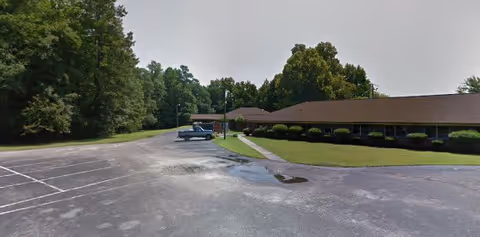 A parking lot adjacent to a single-story building with a brown roof and beige walls, surrounded by green grass and trees under a clear sky.