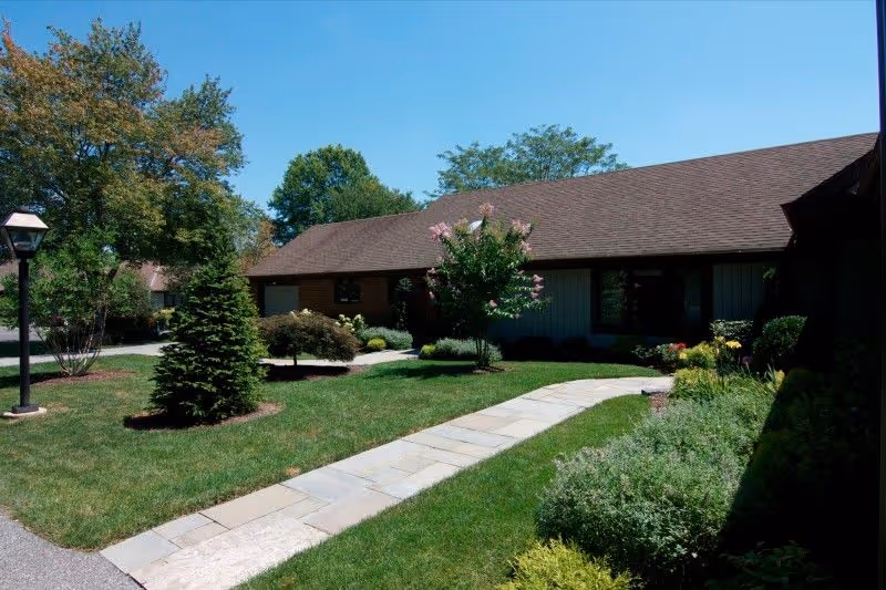 Single-story building with a shingled roof, landscaped lawn, and a stone walkway under a clear blue sky.