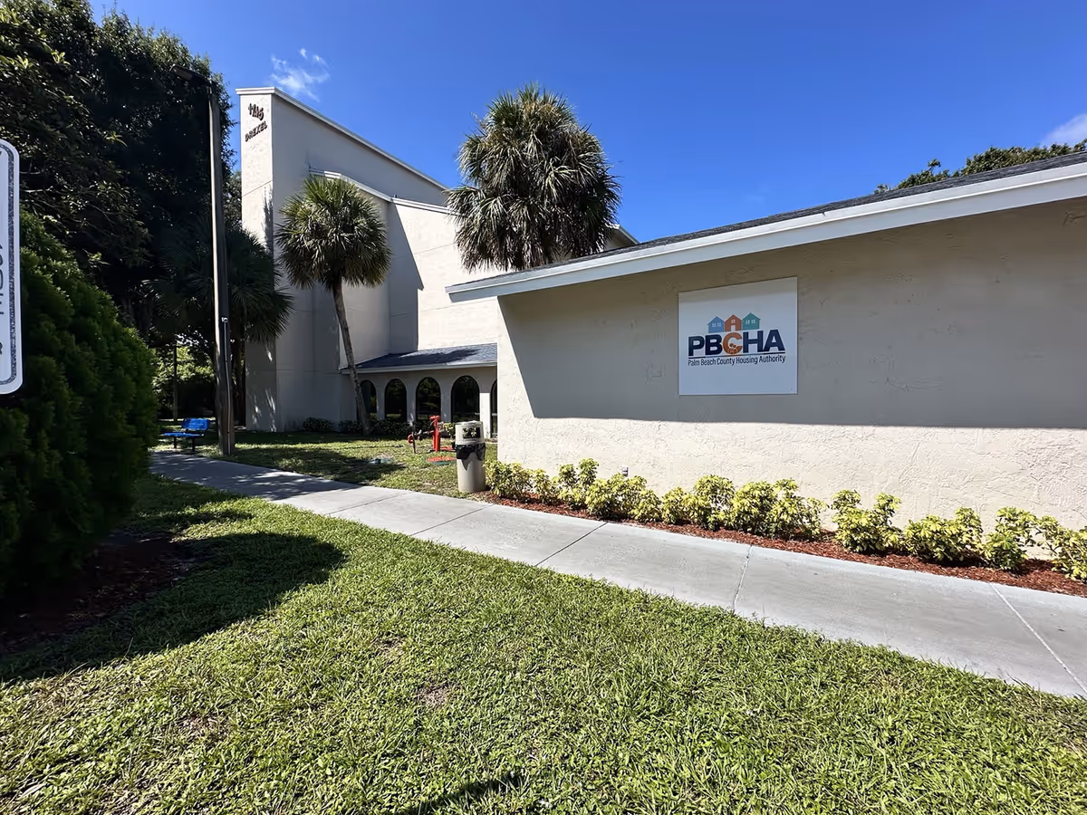 Exterior view of a beige apartment building with a PBCHA sign, palm trees, and a sidewalk.