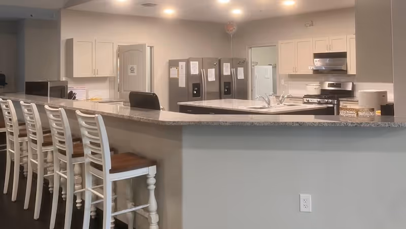 A modern kitchen area with a long granite countertop and six white wooden bar stools with brown seats. The kitchen features white cabinets, two stainless steel refrigerators, a stove with a range hood, a sink, and various kitchen items on the counters. The walls are painted light gray, and the floor is dark wood.