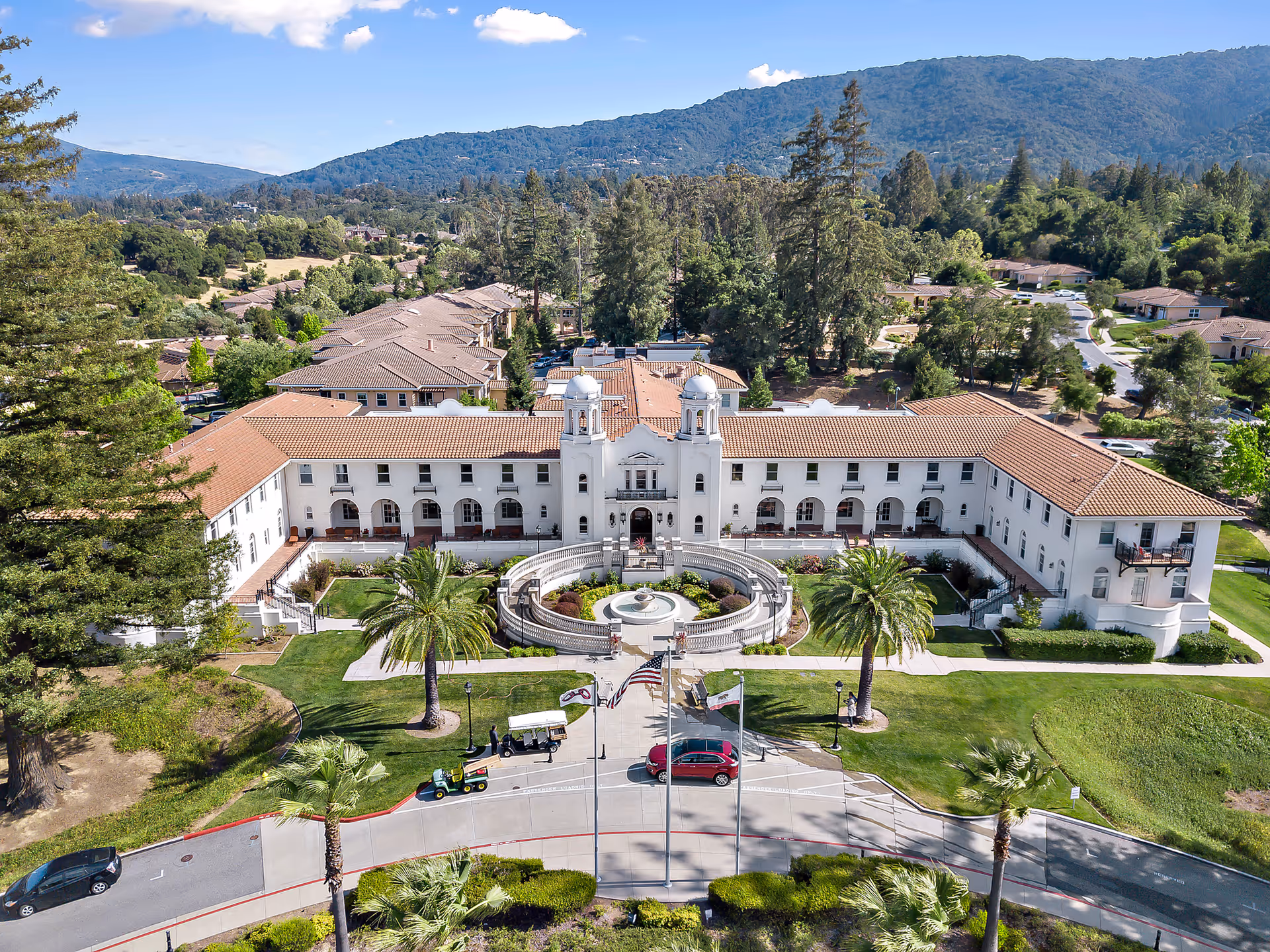Aerial view of a large Mediterranean-style assisted living building with a central circular fountain, palm trees, and surrounding hills.