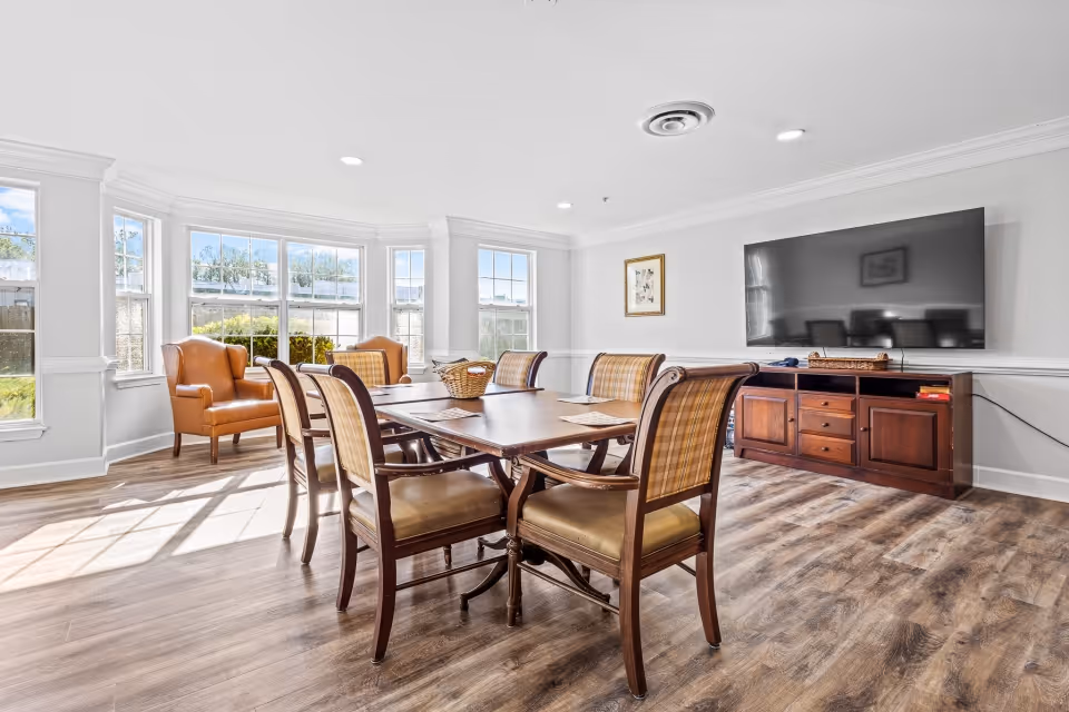 A bright and spacious common area in a senior living facility with a wooden dining table surrounded by six upholstered chairs. Two leather armchairs are positioned near large windows that let in natural light. A flat-screen TV is mounted on the wall above a wooden cabinet. The room has light-colored walls and wood flooring.