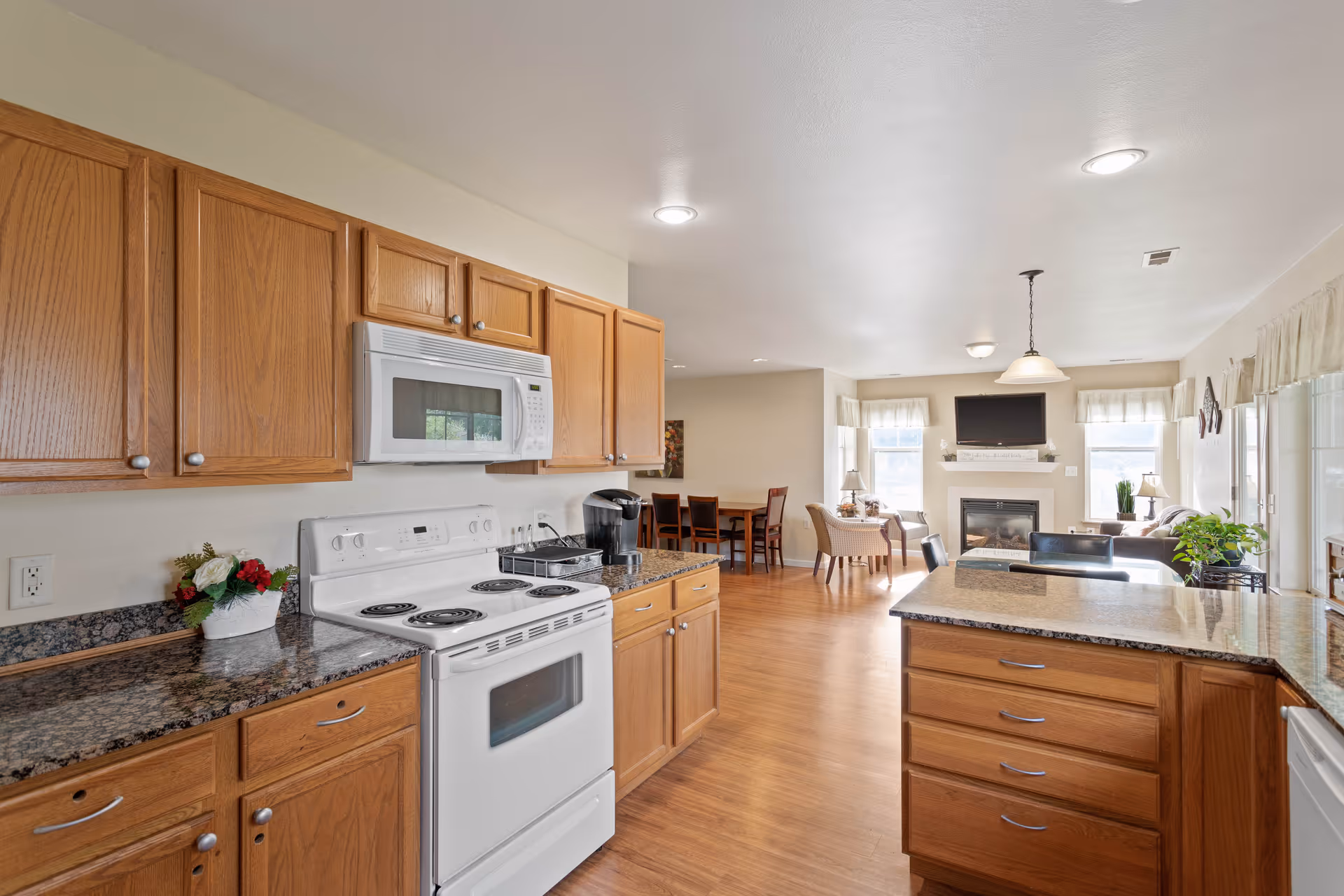 A bright and spacious kitchen area with wooden cabinets, granite countertops, a white electric stove with oven, and a microwave above it. The kitchen opens into a living room with a fireplace, a wall-mounted TV, and seating areas including chairs and a dining table. The room has wooden flooring and large windows letting in natural light.