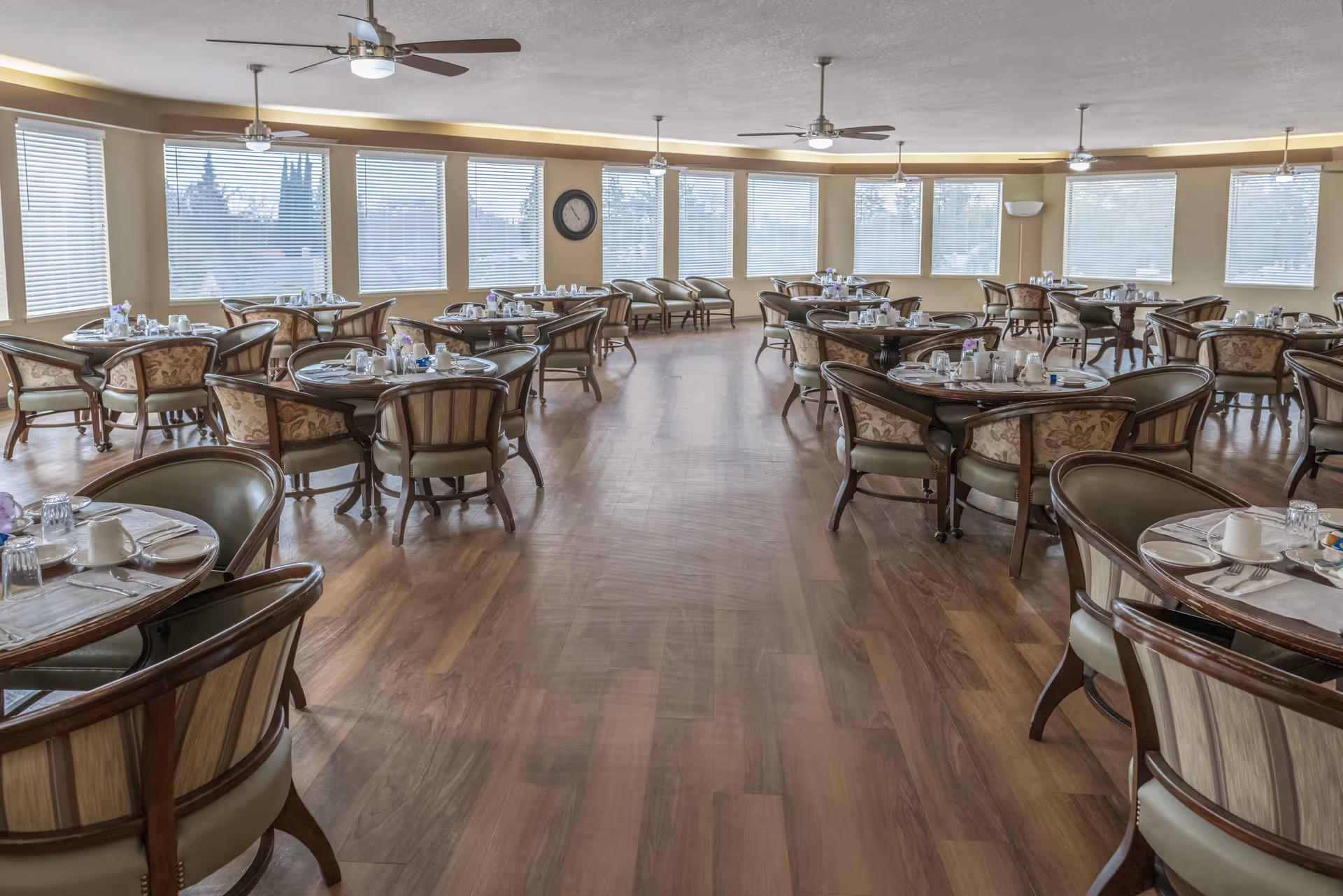 Spacious dining room with multiple round tables set for meals, upholstered chairs, ceiling fans, and large windows along the perimeter.