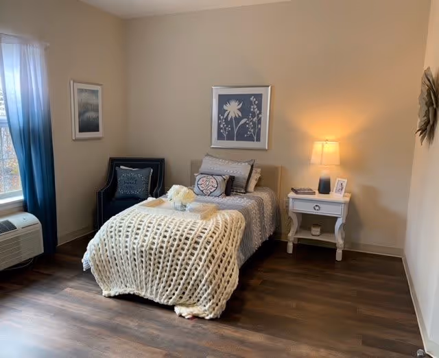 A cozy bedroom with a single bed covered in a chunky knit blanket and decorative pillows. Next to the bed is a white nightstand with a lamp, a framed photo, and a candle. A dark blue armchair with a pillow is placed near a window with blue and white curtains. The room has wooden flooring and neutral-colored walls with framed artwork and a decorative wall piece.
