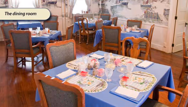 A dining room with several tables covered in blue tablecloths, each set with placemats, napkins, glasses, and condiments. Wooden chairs with cushioned seats surround the tables. The room has wooden flooring and light-colored walls with a mural or wallpaper depicting a waterfront scene.