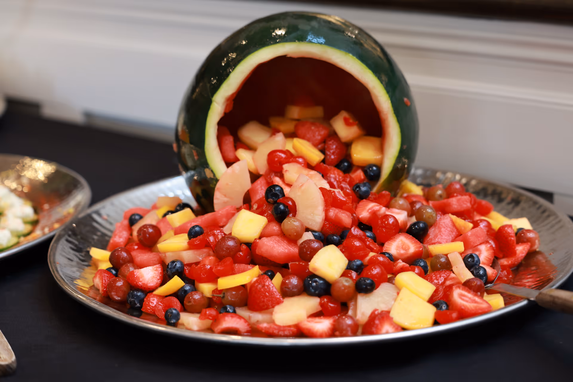 A large silver platter filled with a colorful fruit salad spilling out from a hollowed-out watermelon. The fruit salad contains strawberries, blueberries, grapes, pineapple, and other mixed fruits.