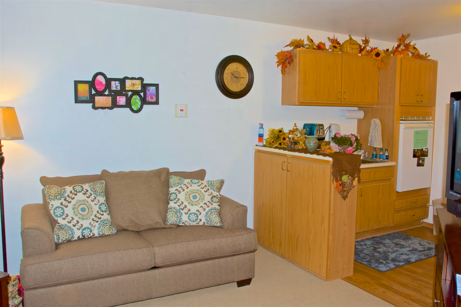 Small living area with a brown sofa and patterned pillows beside a compact kitchenette with wood cabinets and seasonal decorations.