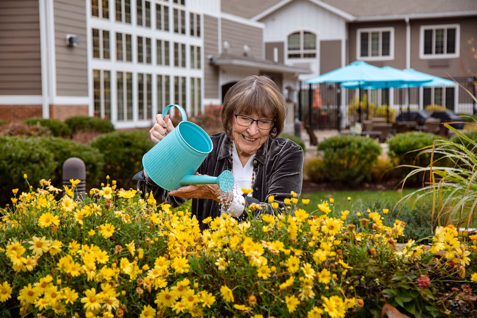 An elderly woman with glasses smiling while watering yellow flowers in a garden outside a senior living facility building with large windows and patio umbrellas in the background.