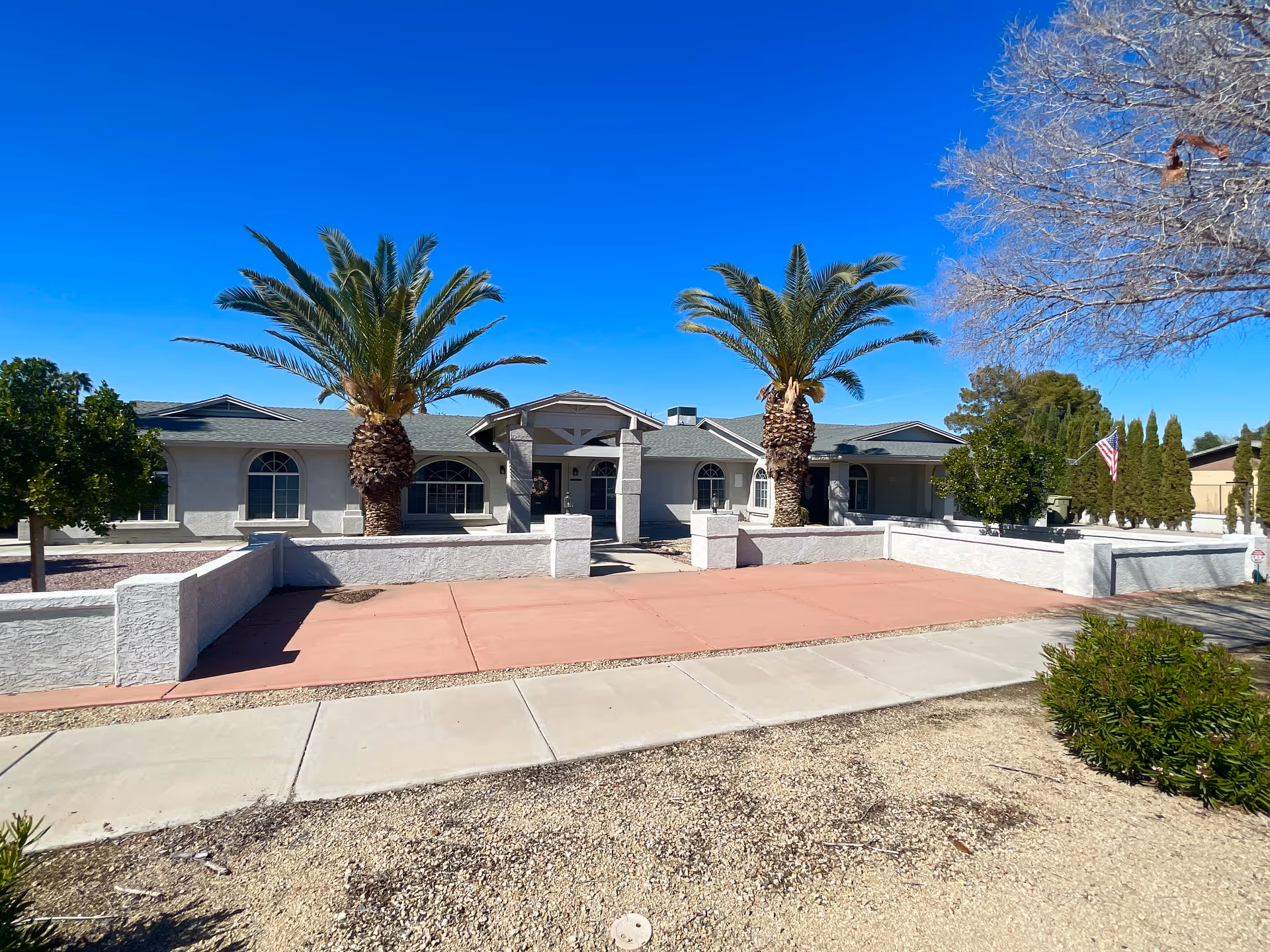 Front exterior view of a single-story building with a light gray facade, green roof, and arched windows. Two tall palm trees are positioned symmetrically in front of the entrance, which has a covered porch supported by columns. The area in front of the building has a pinkish concrete walkway and a low white wall enclosing the front yard. There are additional trees and shrubs around the property under a clear blue sky.