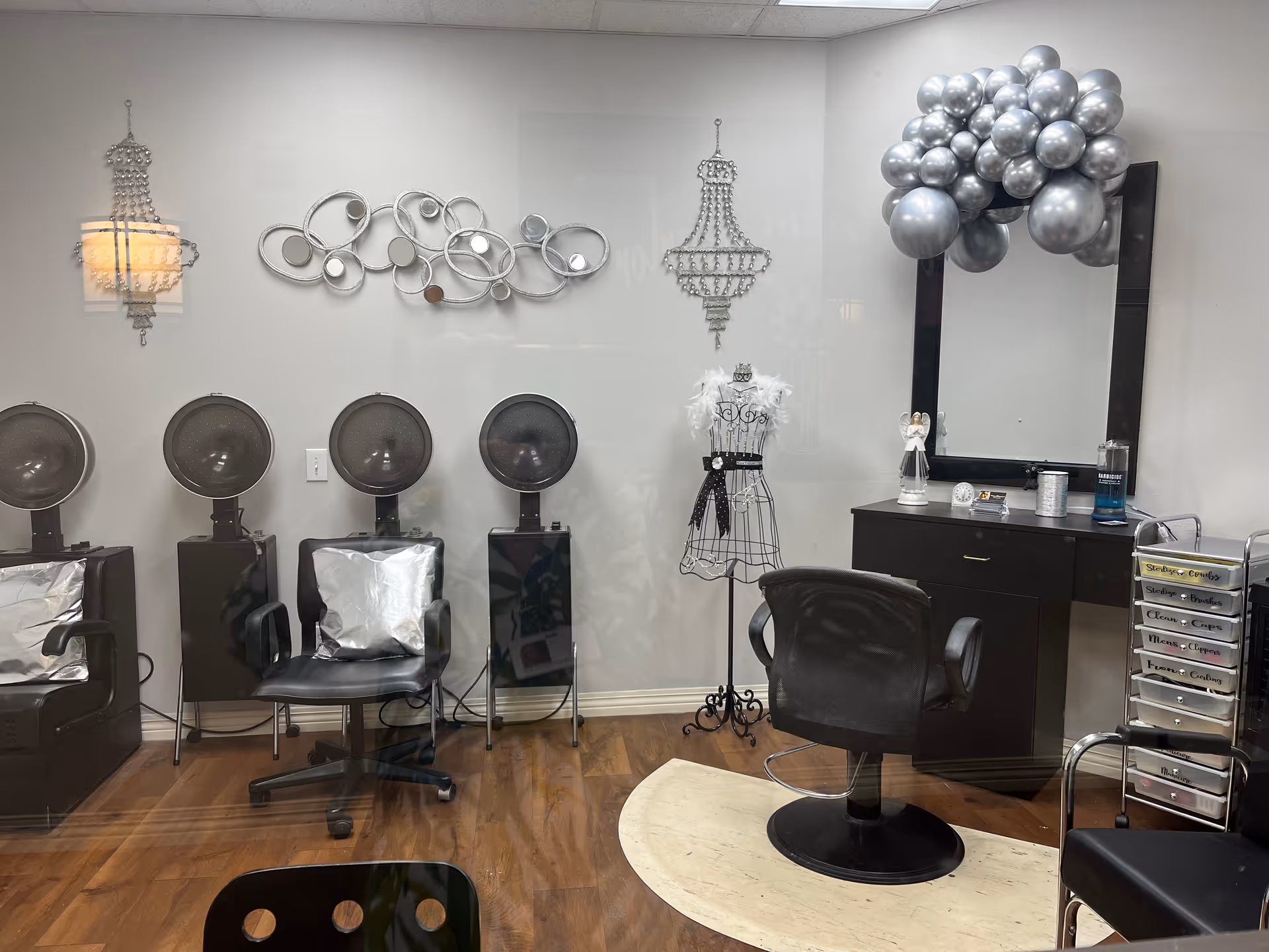 Interior view of a small salon with hooded hair dryers, styling chairs, a mirror and a vanity decorated with silver balloons.